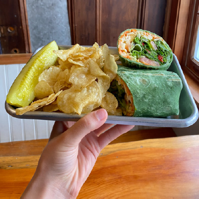A person holding a tray with a wrap, chips and a pickle